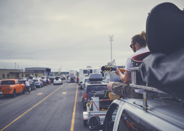 A ponytailed man in his twenties sits on top of a van and plays guitar while waiting to board a Marine Atlantic ferry. 